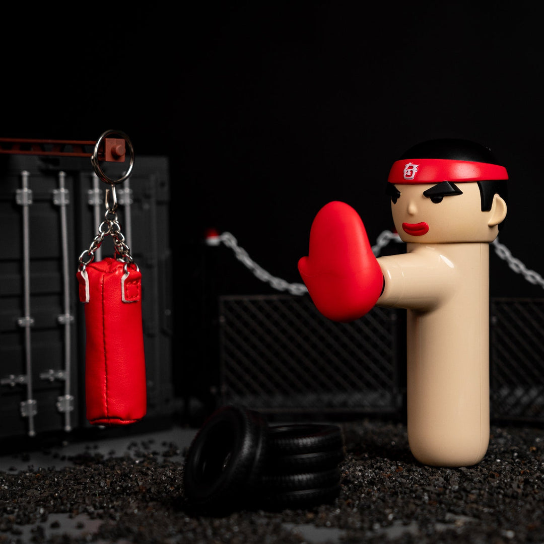 ACECOOL Handmassagegerät KungFu Boxer in beige beim Training mit einem roten Boxsack im Miniatur-Design, Fitness-Setting mit Reifen im Vordergrund.